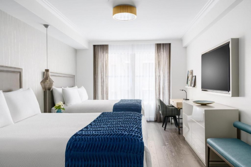 The room has 2 queen-sized beds with a nightstand in between. Opposite the beds is a bench, chest of drawers with a wall-mounted TV above, and a workstation with movable chair.