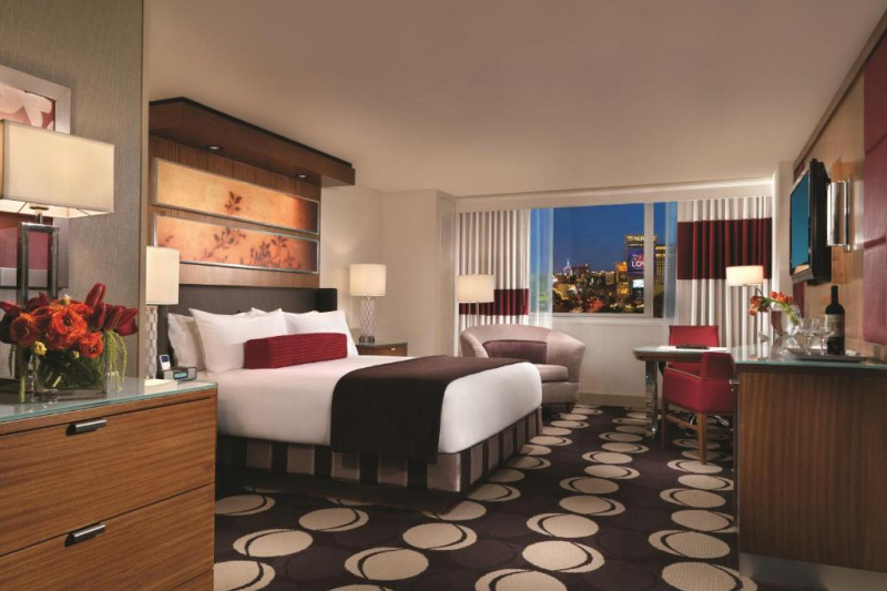 The spacious room has a king-sized bed with nightstands, an armchair, table and chair, desk with executive chair, a wall-mounted TV, and a chest of drawers.
