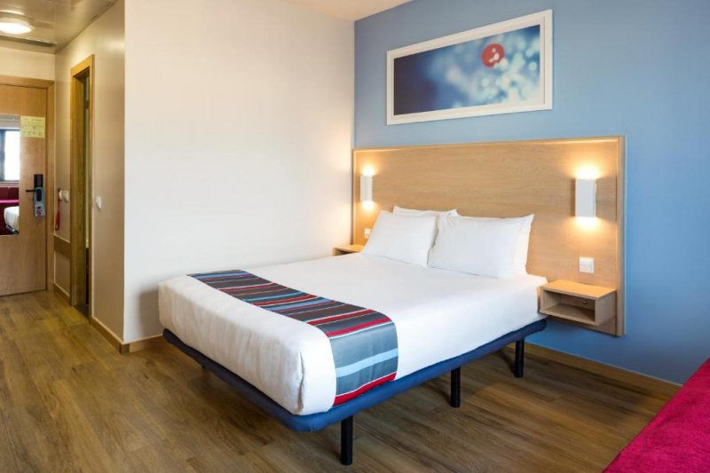 The room has a queen-sized bed with nightstands and a mirror on the room door. There is wooden flooring.