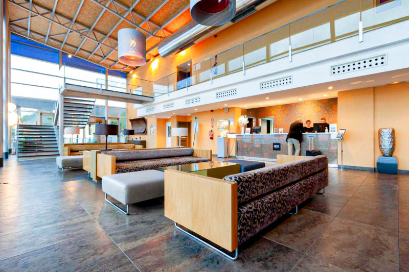 The spacious reception area has a check-in desk with a high counter. There is a seating area with sofas and low tables, and stairs leading to the next floor.