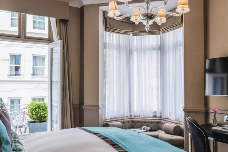 The room has a queen-sized bed, a plush, fitted window seat, a desk with movable chair and a wall-mounted TV. French windows lead to a balcony with a small table and a planter.