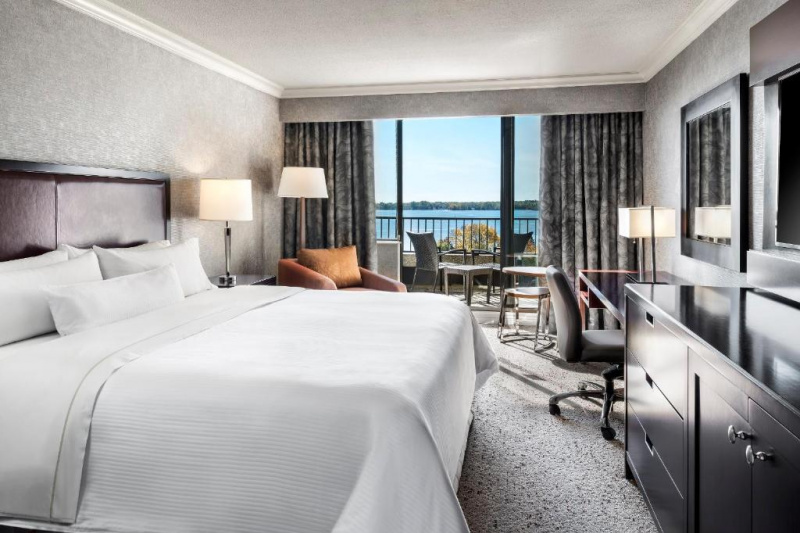The carpeted room has a king-sized bed with a workstation and chest of drawers opposite. To one side are an armchair and table and sliding doors give access to a balcony. Floor space is limited.