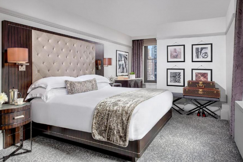 The room has a king-sized bed with nightstands on either side. There is a luggage rack against a wall and a cabinet in the window alcove.