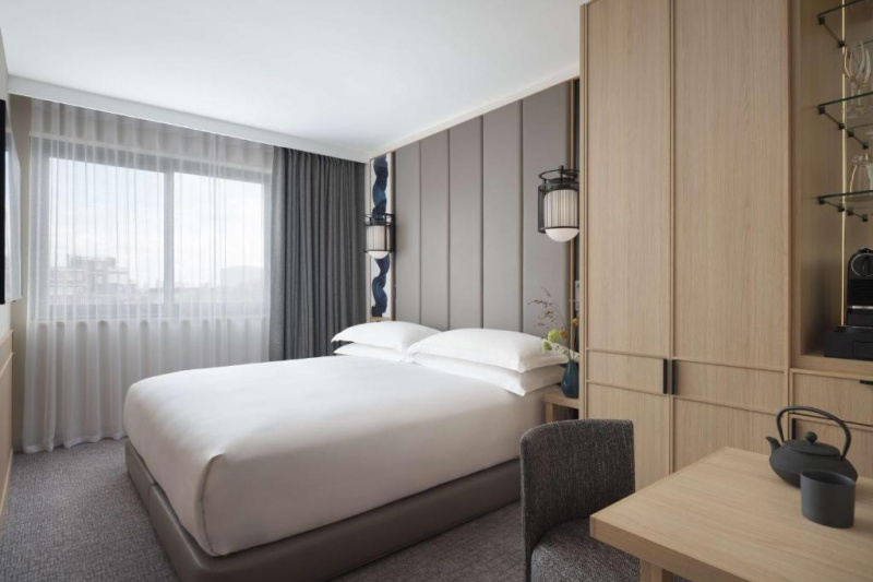 The room has a king-sized bed with nightstands and a wall-mounted TV opposite. There is a small table and movable chair in front of a built-in closet.