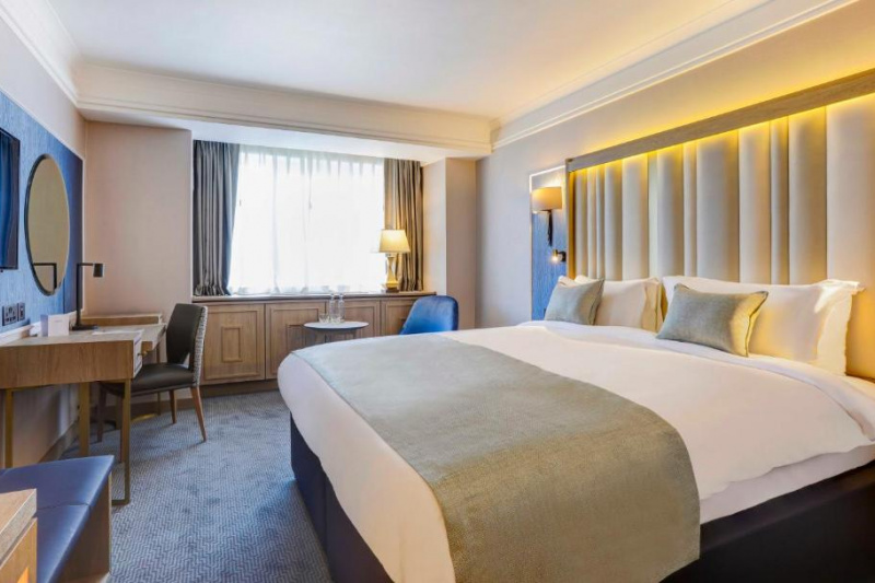 The room has a full-sized bed with nightstands, a workstation with movable chair, a wall-mounted TV, and a chair with side table.