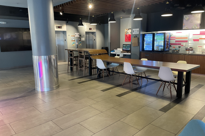 The Re:fuel pantry area in the hotel lobby has a long table with a high section with bar stools and a low section with movable chairs. There is an accessible counter with a drinks refrigerator and snacks, a workstation with a computer and printer and 2 elevators.