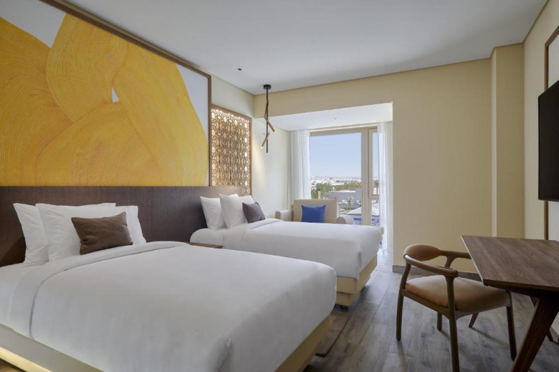 The room has twin beds with a central nightstand, an armchair, a workstation, a chair, and a wall-mounted TV. Floor space is limited.