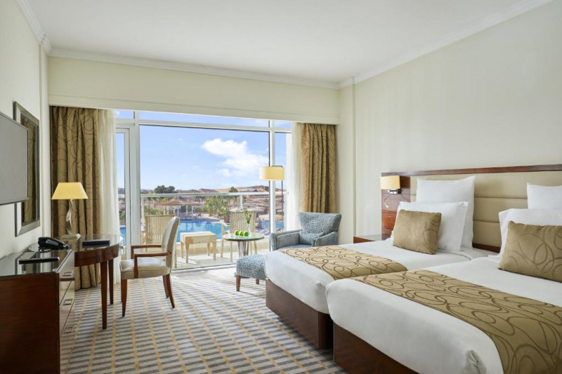 This spacious room has 2 separate beds with nightstands. There is a workstation with a movable chair, a wall-mounted TV, and an armchair with a pouf. The furnished balcony overlooks the pool.