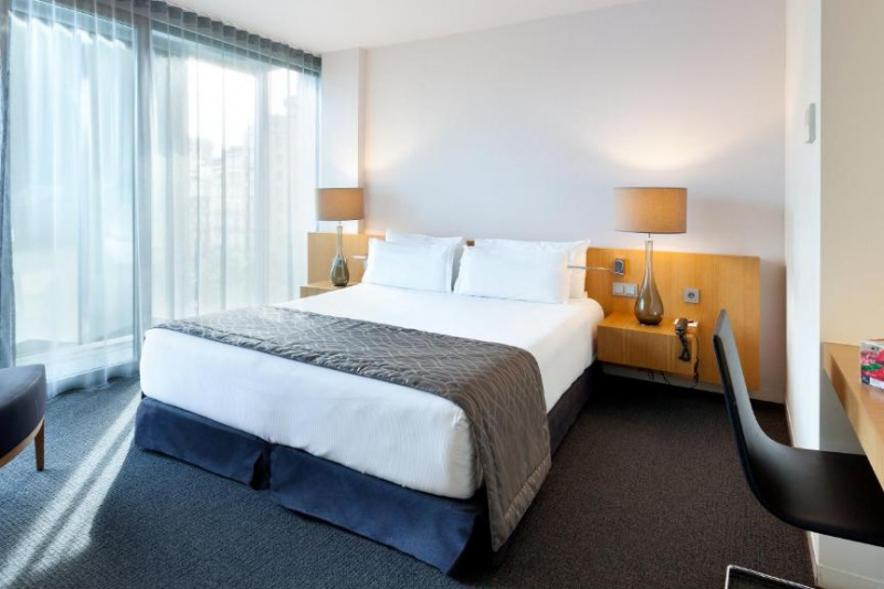 The carpeted room has a full-sized bed with nightstands. There is a built-in workstation with a movable chair, floor-to-ceiling windows and an armchair.