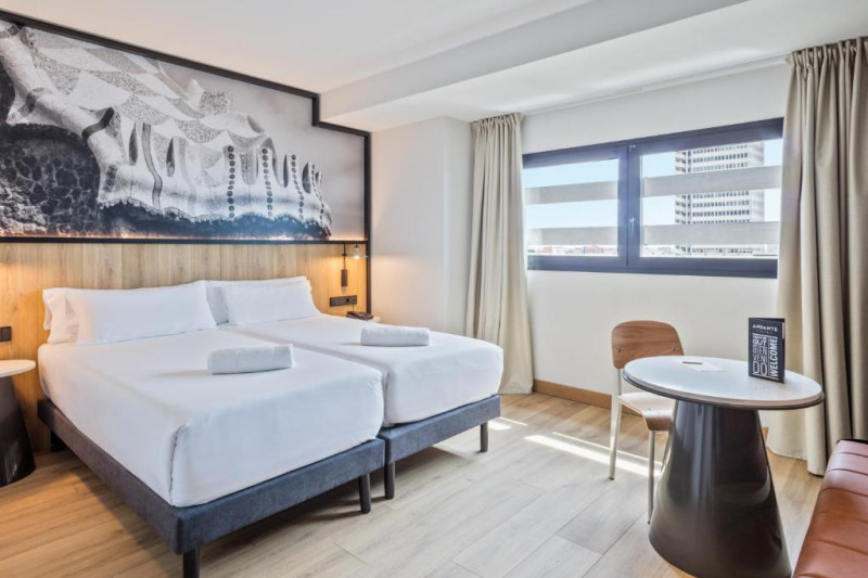 The spacious room has twin beds pushed together with nightstands on either side. There is a small round table, a movable chair and a padded bench.