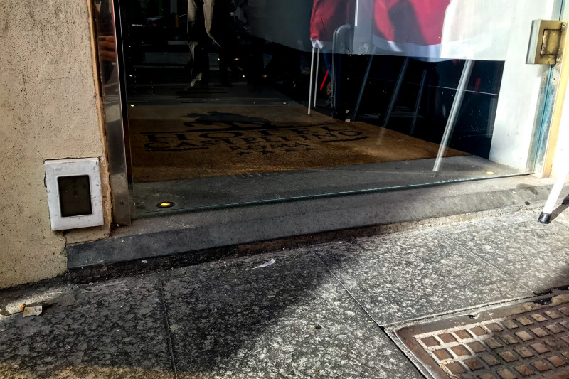 Access to the hotel is through a manual glass door. The entrance is narrow. The entrance is from the sidewalk and there is a small step.