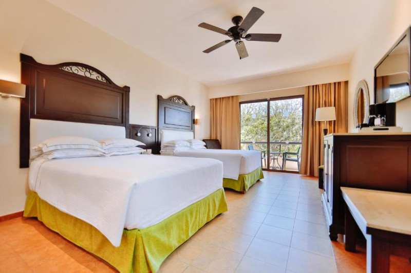 The spacious room has 2 full-sized beds with a central nightstand. There is a wall-mounted TV, a mirror, a cabinet, and an electric fan.