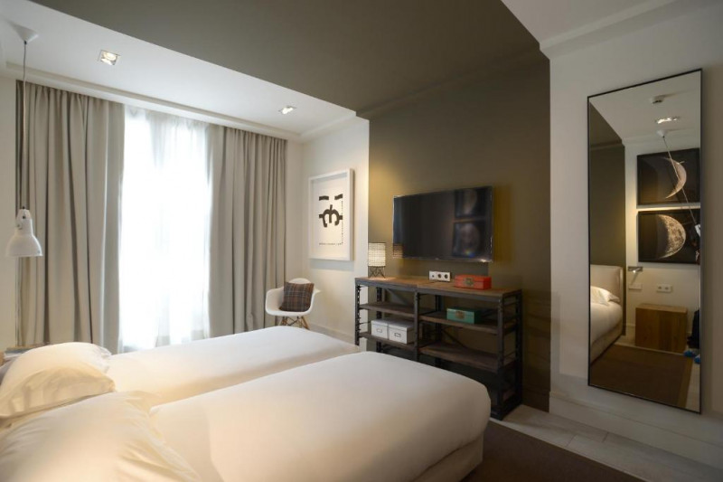 The room has twin beds with nightstands. There is a wall-mounted TV, an armchair,  a shelving unit and a long mirror.