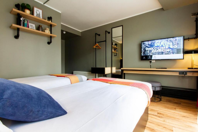 The room has twin beds, a wall-mounted TV, a long workstation and a stool. There is plenty of floor space.