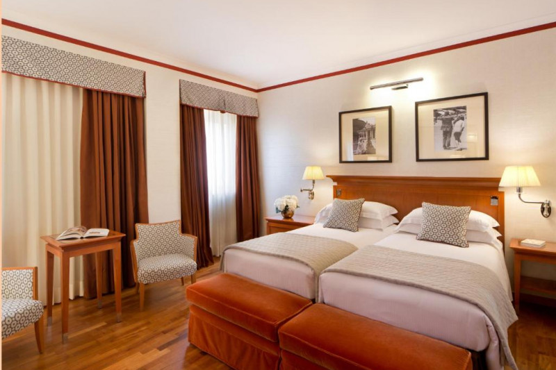 The spacious room has twin beds pushed together with nightstands. There are 2 plush bed benches, 2 armchairs, and a small table.