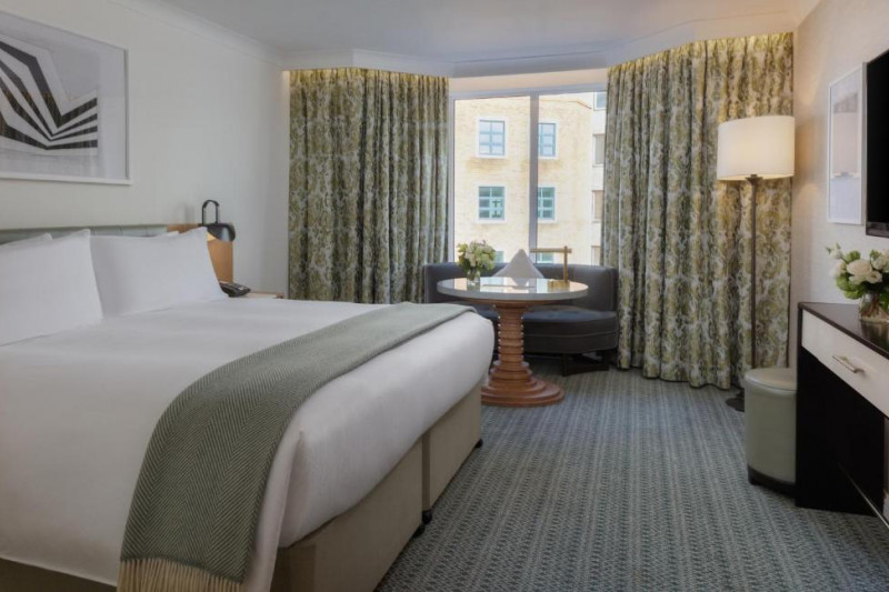 The room has 1 king-sized bed with nightstands. There is a curved sofa in front of the window, a table, a wall-mounted TV and a pouffe.