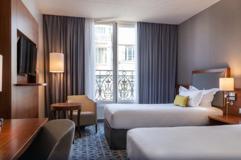 The room has two twin beds with a central nightstand, a workstation, small table, 2 movable chairs, and a wall-mounted TV. Large windows open onto a Juliet balcony.