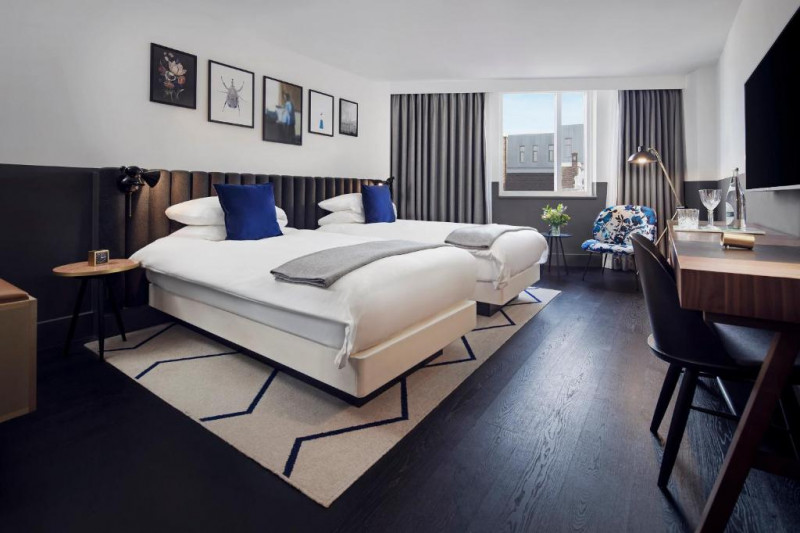 The spacious room has two twin beds with nightstands. There is a workstation, 2 movable chairs, and a wall-mounted TV.