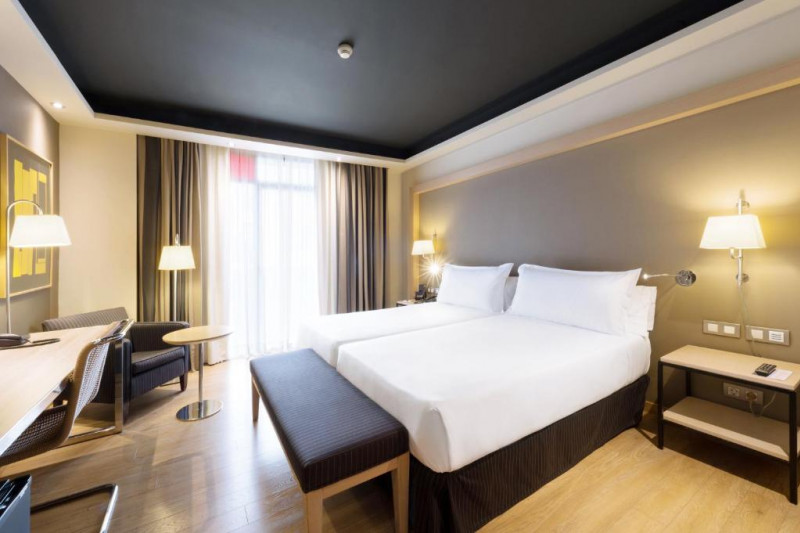 The room has twin beds with nightstands. There is an armchair, a workstation, a chair, and a plush bed bench.