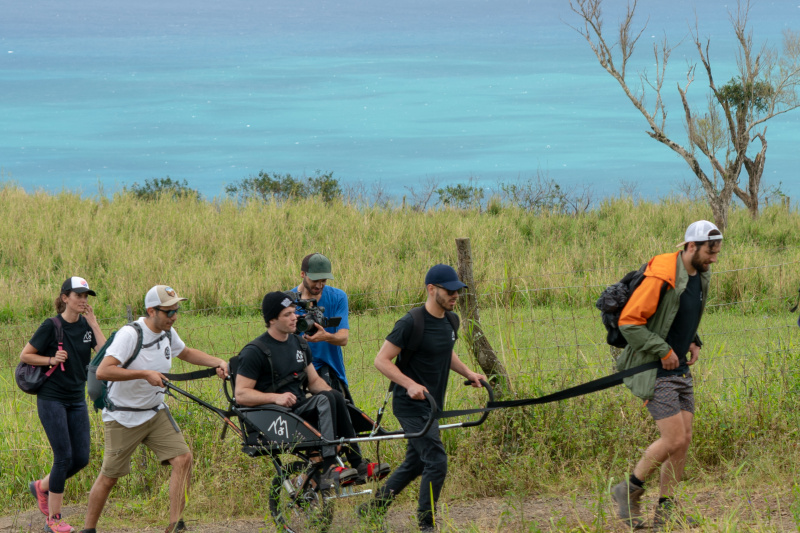 A group of friends travers uneven lands using a joëlette wheelchair, one of them sits on it, and two of them carry the chair working as a team.