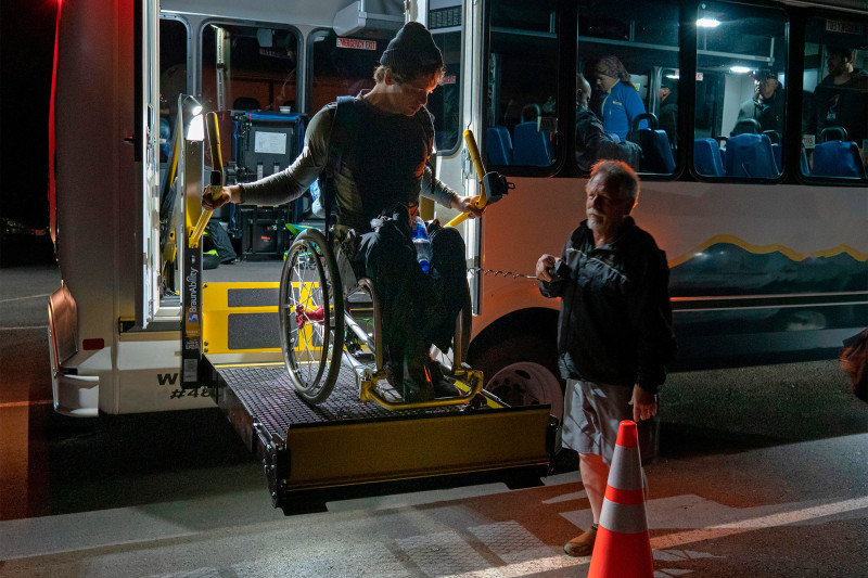 A person in a wheelchair descends from the vehicle via the ramp, with the assistance of a guide.